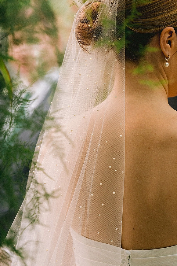 Bridal veil with pearl wedding veil accents draped over a low bun, bride in strapless gown with drop earring, groom blurred in greenery