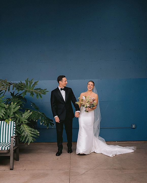 Couple portrait of bride and groom holding hands, laughing by a modern blue wall, bride in veil with bouquet and groom in tuxedo