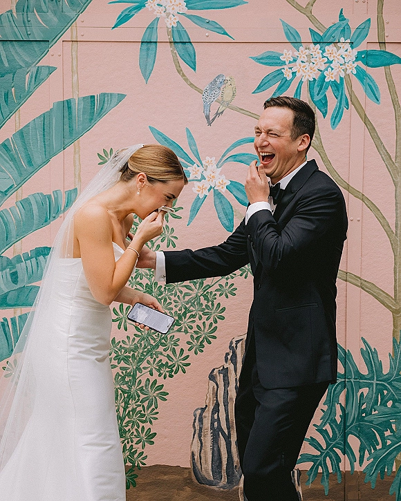 Wedding couple portrait of bride and groom laughing, holding hands in tux and veil, with pearl earrings, before a tropical mural wall