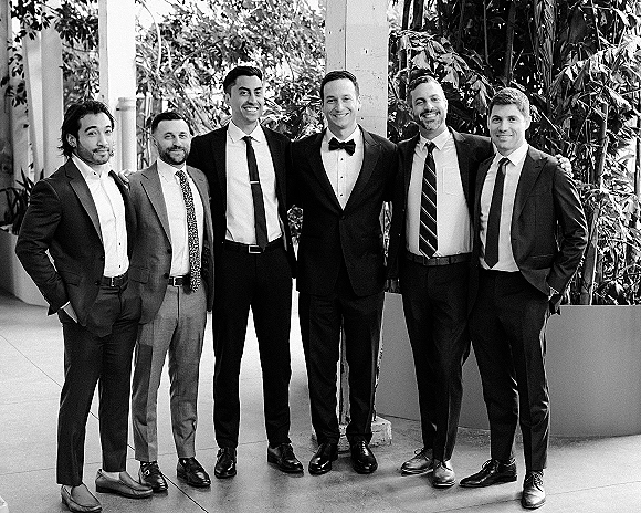 Groomsmen portrait of the groom with groomsmen in suits, posing on an outdoor patio with greenery, potted plants, and columns, bow tie accent