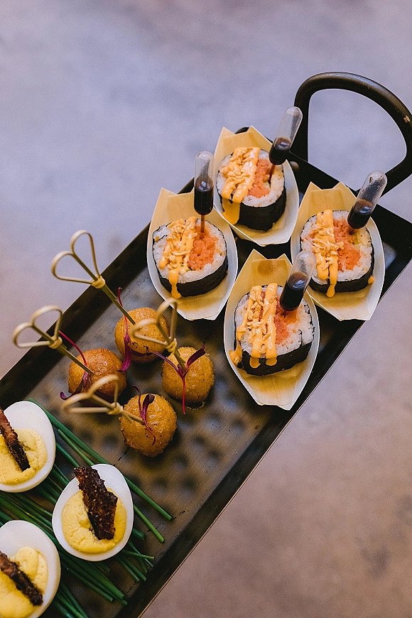 Wedding cocktail appetizers on a serving tray with sushi bites, deviled eggs, fried skewers, and sauce pipettes on a concrete floor