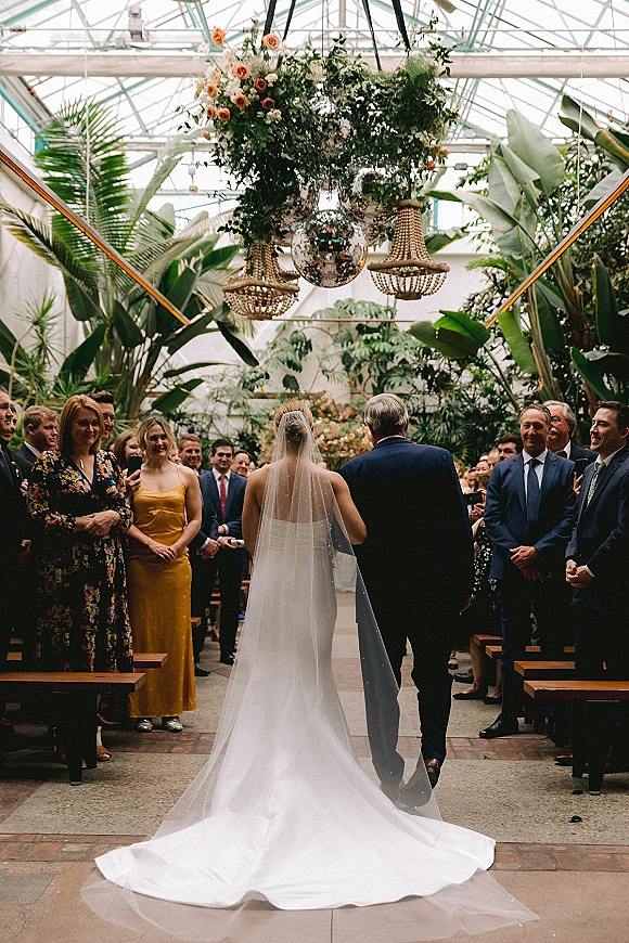 Wedding processional with bride walking down aisle from behind, veil and long train under floral chandelier in tropical greenhouse venue