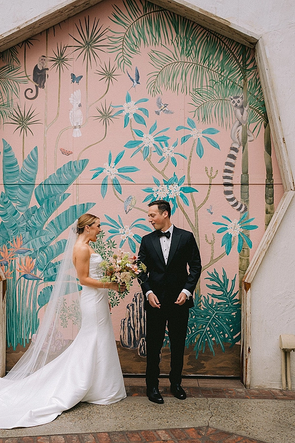 Couple portrait of bride in veil holding a bouquet beside groom in black tuxedo and bow tie, posed by a colorful tropical mural wall