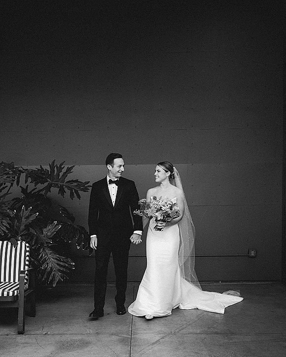 Couple portrait of bride and groom holding hands, bride with bouquet and veil, in front of plain wall with tropical plants