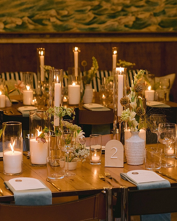 Reception tablescape with candlelit wedding table styling, glass hurricane candles and bud vase florals on a long wooden table in a dim room with mural backdrop
