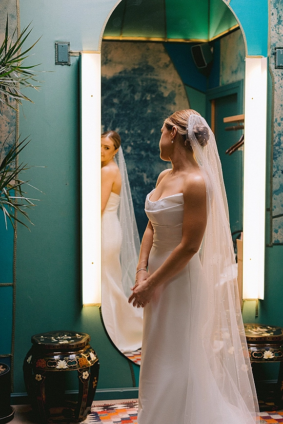 Bridal portrait with bride in mirror wearing a strapless wedding dress and cathedral veil, pearl earrings, against a teal wall archway