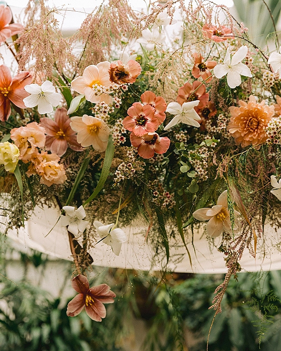 Wedding floral arrangement with peach and white wildflowers and trailing greenery, softly set against blurred garden foliage outdoors