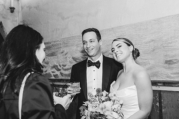 Wedding reception moment in black and white as bride with bouquet and groom in tuxedo laugh with cocktails by a mural wall sconce