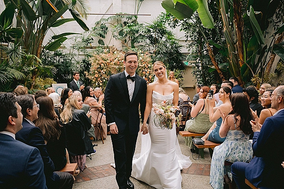 Wedding recessional as bride and groom walk the aisle, her bouquet and veil flowing, guests applauding in an indoor garden greenhouse setting