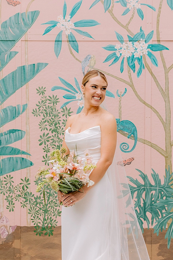 Bridal portrait of a bride in a strapless satin wedding dress, smiling with bouquet and veil before a pink tropical mural wall