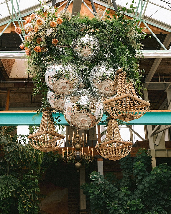 Wedding chandelier decor with disco ball wedding decor, lush greenery and orange florals, hanging under a glass ceiling with metal beams