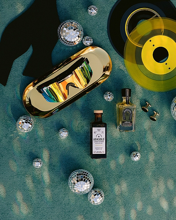 Groom accessories flat lay featuring a black bow tie, mirrored sunglasses, cufflinks, cologne, liquor bottles and disco balls on teal fabric