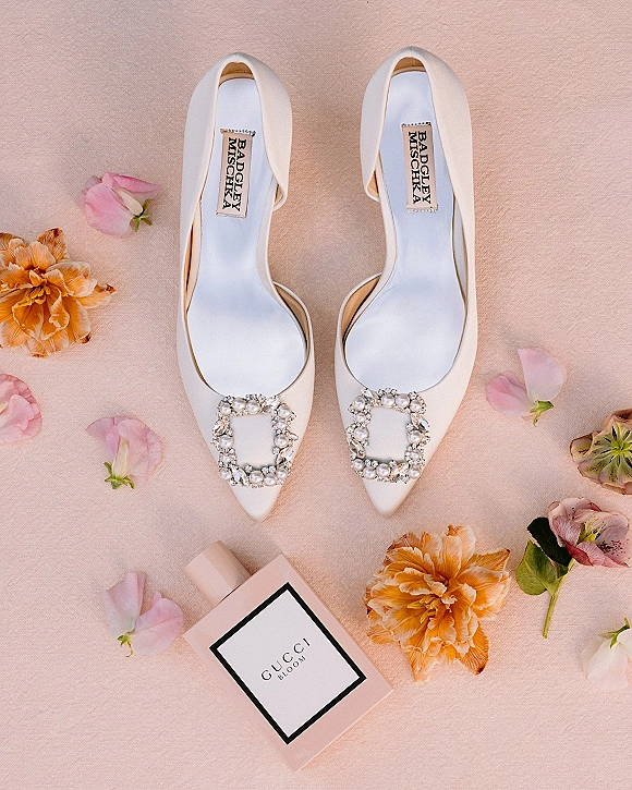 Bridal shoes flatlay with white pointed-toe heels, rhinestone buckles, and perfume bottle on blush fabric scattered with rose petals