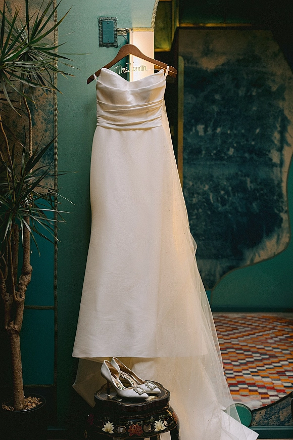 Wedding dress hanging on a wooden dress hanger beside bridal shoes with jeweled buckles, set against a teal wall with framed art