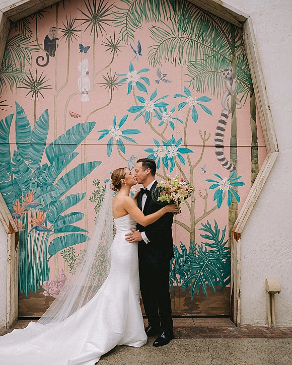 Wedding kiss portrait of bride and groom kissing, her cathedral veil and bouquet flowing beside a colorful tropical mural wall doorway backdrop