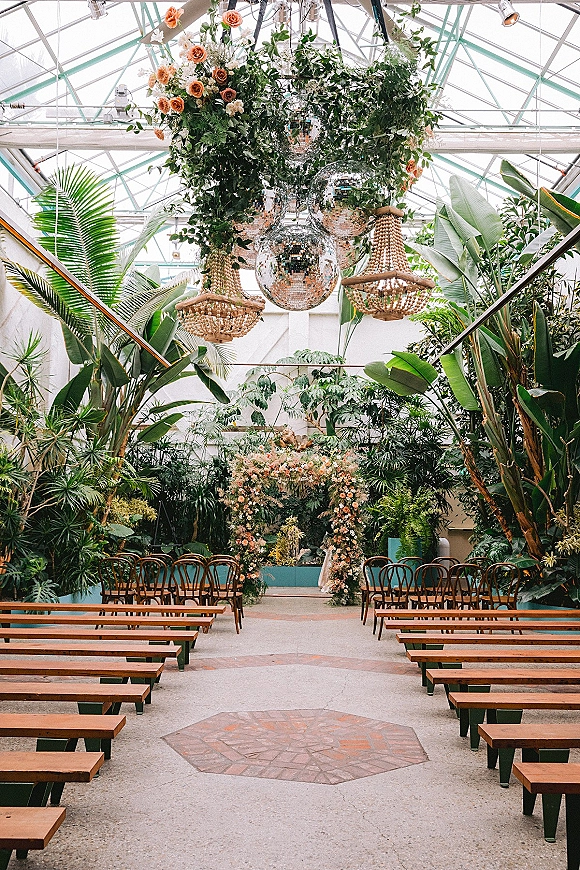Ceremony setup in a greenhouse wedding ceremony with a floral arch, hanging greenery, disco balls, and wooden benches under glass ceilings