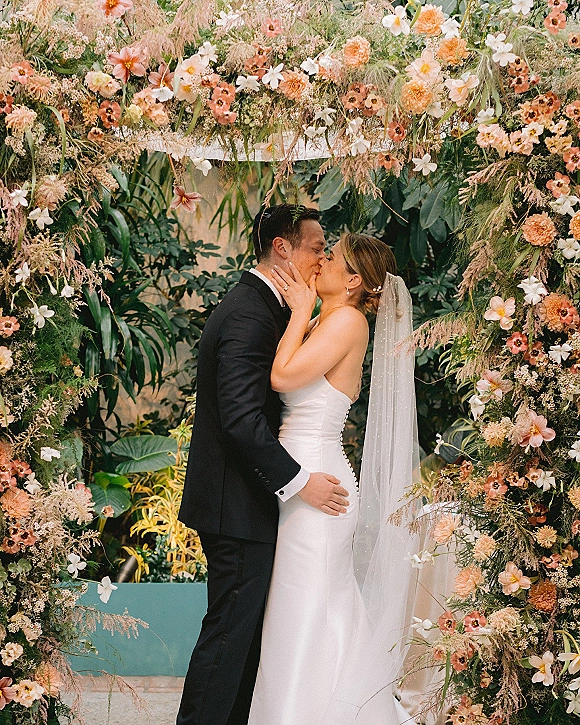 Wedding kiss portrait of bride in strapless dress and veil holding groom’s face in tuxedo beneath a floral arch in indoor garden greenery