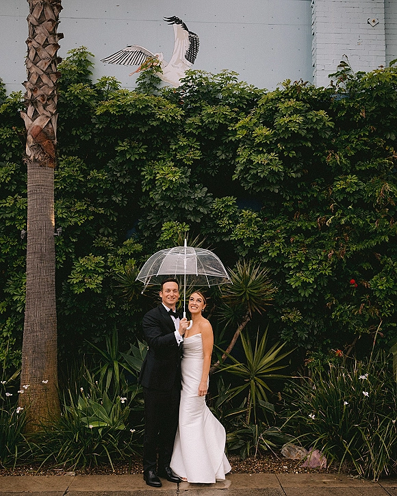 Couple portrait of bride in strapless wedding dress and groom in tuxedo posing under a clear umbrella beside lush palms and mural wall