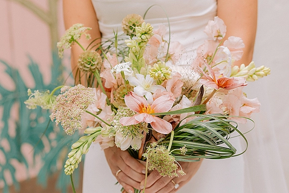 Bridal bouquet of blush and peach flowers with white blooms and greenery, held against a strapless gown with an engagement ring on display