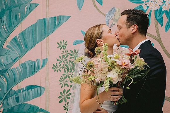 Wedding kiss portrait of bride and groom kiss, bride holding bouquet with veil and earrings against a pink palm-leaf mural wall