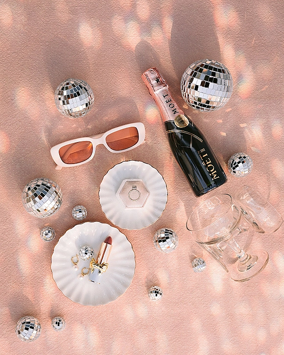 Wedding detail flatlay with engagement ring, champagne bottle, coupe glasses, and mini disco balls on pink fabric with light reflections