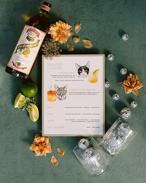 Signature cocktail menu with wedding cocktail menu card styled beside limes, cut glass tumblers, and disco balls on green fabric backdrop
