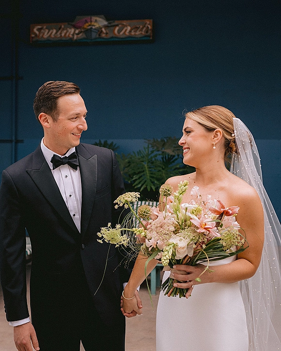 Couple portrait of bride and groom holding hands, looking at each other, with colorful bouquet and veil against a blue wall with greenery