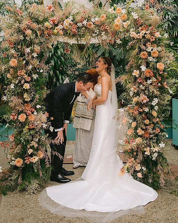 Ceremony moment as bride and groom laugh with officiant under a floral arch, in a greenhouse with plants, teal wall, and veil flow