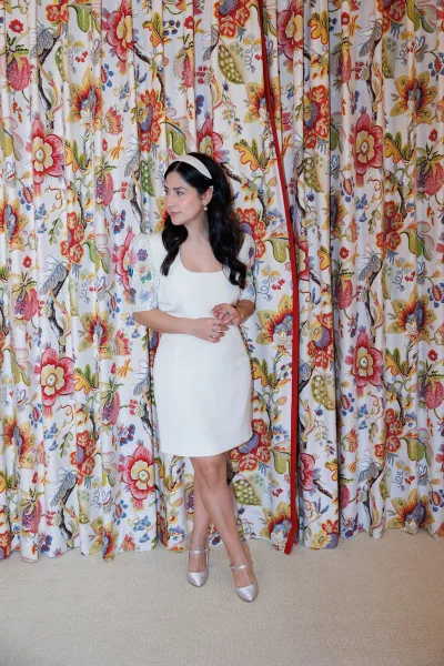 Bridal portrait of a bride in a short wedding dress with embroidered sleeves, posing before a floral curtain on a carpeted floor.