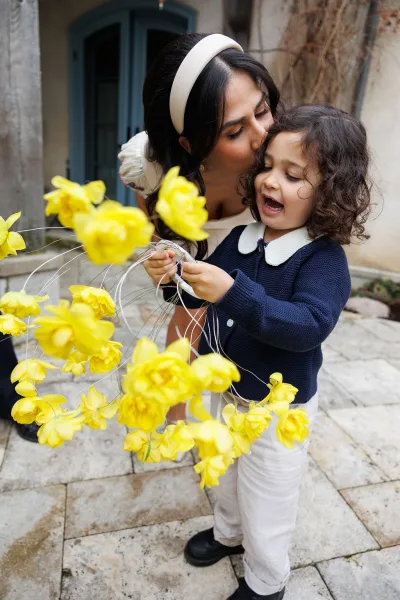 Wedding candid moment as a bride kisses a child in a navy cardigan on a stone patio, with a blue door and vine-clad stucco wall behind.