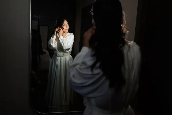 Bride getting ready, bride putting on earrings by a mirror in a dark room, pausing near a doorway as soft light outlines her calm profile.