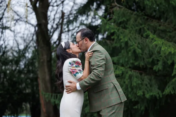 Wedding kiss portrait where a bride and groom embrace, her birdcage veil brushing his cheek as they kiss amid evergreen trees in a quiet forest.