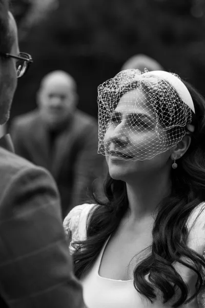 Ceremony moment with a bride in a birdcage veil smiling toward her groom, the short vintage veil framing her face in a tender, candid exchange.