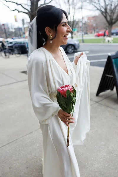 Bridal portrait of a bride holding protea on a city sidewalk, framed by leafless trees, parked cars, and bicycles in the background.