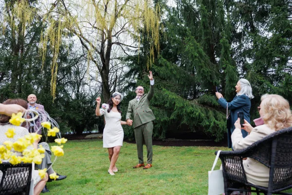 Wedding recessional during a just married exit, the couple walks the garden lawn aisle as guests wave floral wands beneath willow branches.