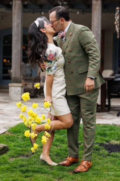 Wedding kiss portrait with a bride lifting her leg in a playful pose, holding a yellow bouquet on a stone patio beneath string lights.