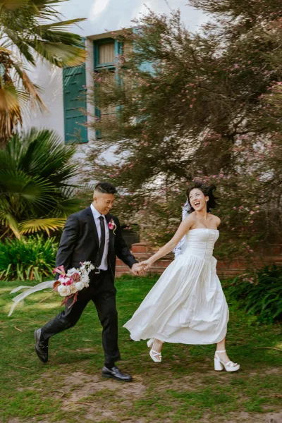 Couple portrait of bride and groom running hand in hand, groom holding bouquet, bride laughing in veil on palm-lined garden lawn