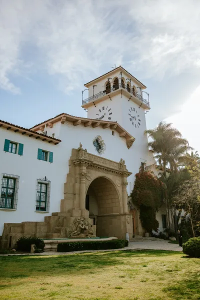 Wedding venue exterior with a clock tower above an arched entryway, white stucco walls and clay tile roof, framed by palms and a fountain courtyard lawn