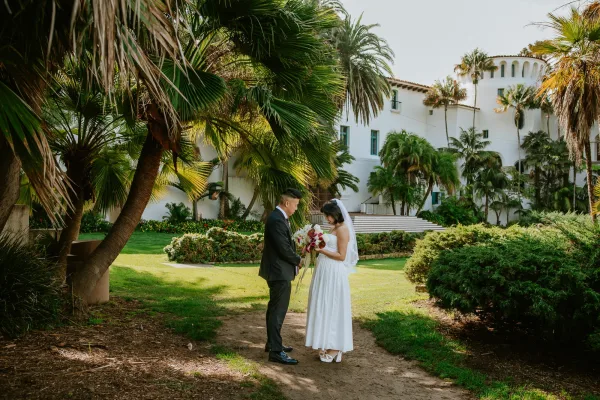 Couple portrait at an outdoor wedding, bride in a strapless dress holding a colorful bouquet beside groom in black suit under palm trees