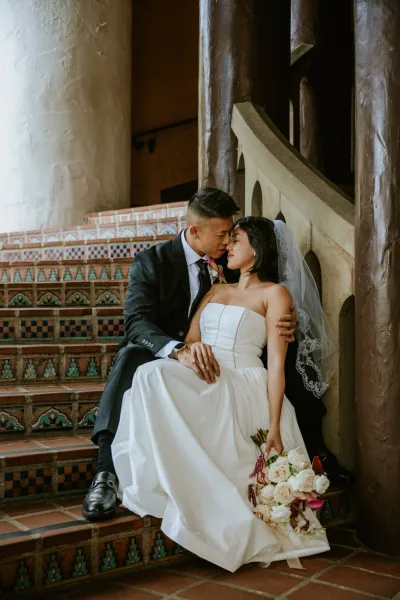 Wedding couple portrait of bride and groom sitting on stairs, foreheads touching as she holds a white rose bouquet on tiled steps