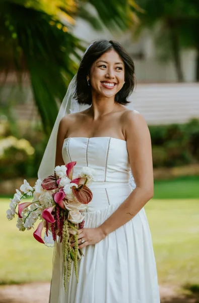 Bridal portrait of a smiling bride holding bouquet in a strapless satin dress and veil, standing by palm leaves on a garden walkway