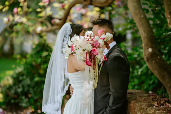 Wedding kiss as bride and groom kiss behind a pink-and-white orchid bouquet, her veil draped softly in a garden with stone wall backdrop