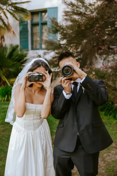 Couple portrait of bride and groom with cameras covering their faces, her veil and his boutonniere in a garden with palms and shutters