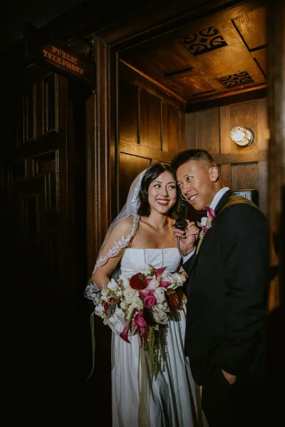 Couple portrait of bride and groom in a phone booth, she holds a calla lily bouquet as he lifts a vintage receiver in moody wood paneling