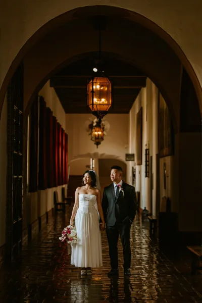 Couple portrait of bride and groom walking hand in hand, her strapless dress and tulip bouquet beneath lantern-lit arched hallway with red curtains