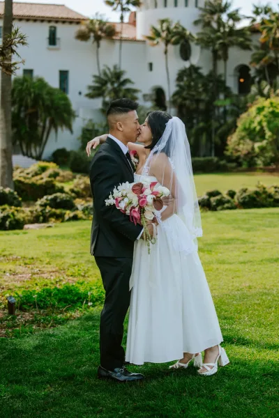 Wedding kiss portrait of bride and groom kissing, bride holding bouquet with veil flowing on a lawn by palm trees and a white stucco venue