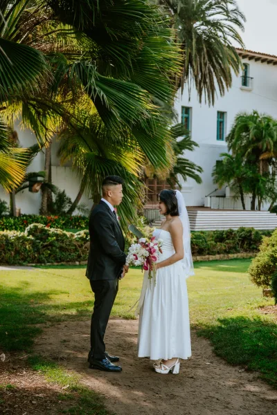 Wedding vows as bride reads from a vow booklet to groom, veil trailing, with palm trees and a white villa garden backdrop in daylight