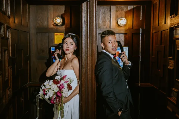Couple portrait in a wood-paneled phone booth, bride with calla lily and orchid bouquet as groom talks on a vintage payphone