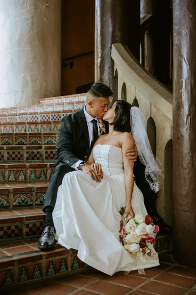Wedding kiss portrait of bride and groom kissing on a tiled staircase, her veil and rose bouquet visible beside his black suit