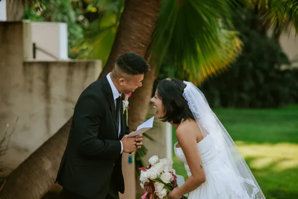 First look moment as bride and groom laughing while he reads vows, her veil and bouquet visible on a garden lawn with palm trees